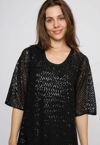 Top negro con lentejuelas, de ajuste holgado, mangas cortas y escote redondeado. Presenta un diseño texturizado y estampado con detalles brillantes.
