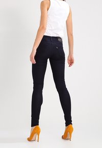 Jeans skinny de mezclilla oscura con cintura alta, que presentan una textura suave y bolsillos traseros mínimos, combinados con zapatos de tacón alto naranjas.