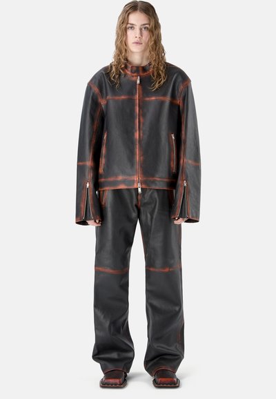 Personne aux longs cheveux bouclés portant une veste en cuir noire et un pantalon avec des surpiqûres rouges et des fermetures éclair, debout devant un fond blanc uni.