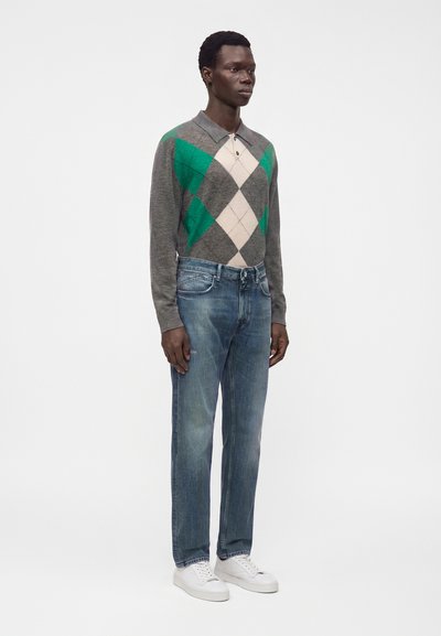Homme debout portant un pull à losanges gris et vert, un jean bleu et des baskets blanches sur un fond blanc uni.