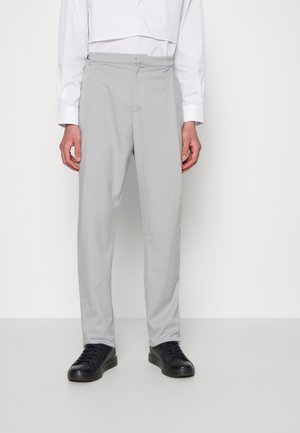 Mężczyzna w jasnoszarych, dopasowanych spodniach, białej koszuli na guziki i czarnych trampkach, stojący na jednolitym białym tle.