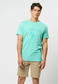 Tricou din bumbac de culoare teal deschis, cu guler rotund și mâneci scurte, având o imprimeu grafic cu un jucător de polo și denumirea brandului în teal închis. O pereche de pantaloni scurți bej.
