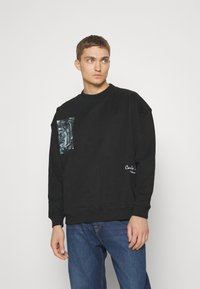 Zwarte sweatshirt met een grafische pocketpatch en geborduurd logo. Ronde hals, lange mouwen, relaxte pasvorm, gemaakt van een zachte katoenmix.