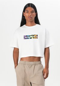 Hvit, avkortet t-skjorte med et fargerikt, abstrakt Converse-logo foran. Stoffet ser glatt ut med innsydd kortermet.