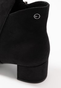 Černá nubuková kotníková bota s robustním podpatkem, vybavená bočním zipem a kulatým kovovým logem na svršku.