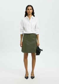 Biała koszula z guzikami z podwiniętymi rękawami, oliwkowa skórzana spódnica i czarna torba; modelka w czarnych szpilkach z ostrym noskiem.