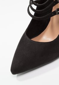 Chaussure à talon haut en daim noir à bout pointu avec doubles brides autour de la cheville et une boucle argentée sur fond blanc.