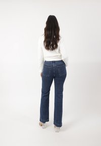 Donkerblauwe denim flare jeans met twee achterzakken, gedragen met een witte langmouwtop. Duidelijke hakken zijn zichtbaar aan de onderkant.