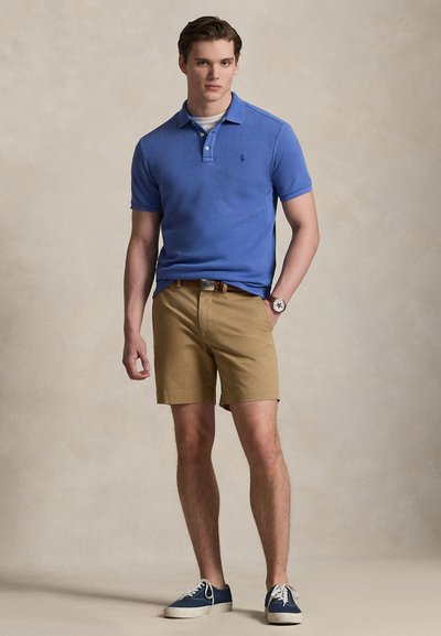 Νέος άνδρας όρθιος, φοράει μπλε πόλο πουκάμισο, καφέ σορτς με ζώνη, μπλε αθλητικά παπούτσια και ρολόι χειρός, ουδέτερο φόντο.