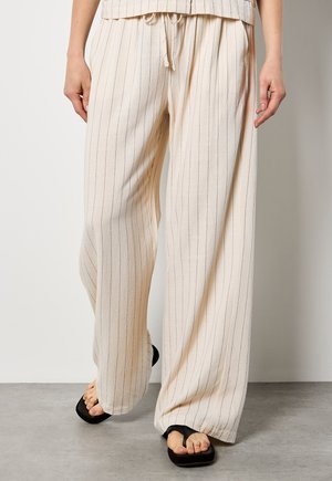 Vrouw draagt crèmekleurige broek met brede pijpen en dunne verticale strepen, gecombineerd met zwarte teenslippers, staand op een grijze vloer tegen een witte muur.