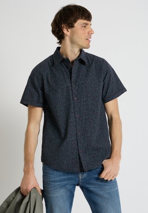 Jonge man die een donker patroon met korte mouwen overhemd en spijkerbroek draagt, houdt een grijze jas in zijn linkerhand.