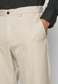 Pantaloni beige in tessuto morbido, con taglio dritto, tasche laterali e un solo bottone nero in vita.