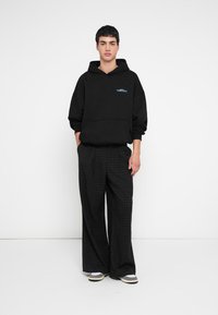 Svart hettegenser med frontlomme, med en liten blå logo, kombinert med løse svarte rutete bukser og tofargede sko.