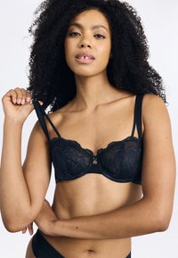 Svart spets-bh med genomskinliga kupor, dekorativa vågiga kanter och en central utsmyckning. Justerbara axelband och ett mjukt band nedanför.