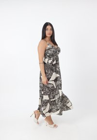 Mujer con cabello largo y oscuro, vestida con un maxi vestido sin mangas de patrón de hojas en blanco y negro y sandalias blancas de tacón, posando frente a un fondo blanco liso.