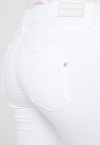 Primer plano de una persona que lleva vaqueros Replay blancos, enfocándose en el bolsillo trasero y la etiqueta de cuero con la marca en la cintura.