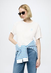 Tricou din bumbac bej deschis cu mâneci scurte și un logo albastru. Combinat cu un hanorac albastru cu dungi albe legat în jurul taliei, blugi din denim.