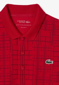 Röd polo skjorta med en strukturerad rutmönster, med klassisk krage, två knappar på placket och en grön krokodil-logotyp som accent. Regular fit.