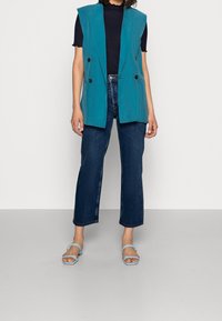 Tyrkysový bezrukávový blejzr s dvojitým zapínáním, nošený přes černý top, v kombinaci s tmavomodrými rovnými džínami a světle modrými sandály.