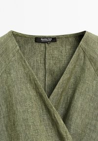Tissu vert olive avec un design en V, portant une étiquette indiquant "Massimo Dutti, fabriqué au Portugal." Matériau en lin texturé visible.