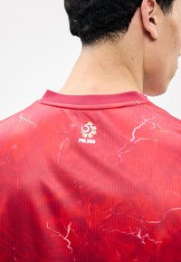 Personne portant un maillot de sport rouge de la Pologne avec des motifs d'éclairs blancs dans le dos, vue de dos et légèrement de côté.