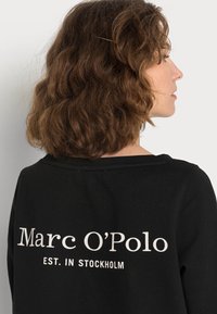 Černá mikina s volným střihem, na zadní straně s bílým nápisem "Marc O'Polo" a "EST. IN STOCKHOLM". Měkký bavlněný materiál.