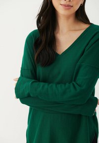 Vihreä V-kaula-aukkoinen neule, jossa on pitkät hihat, valmistettu pehmeästä kankaasta. Ominaisuuksiin kuuluu rento istuvuus ja sivuhalkiot. Pintakuvio vaikuttaa sileältä.