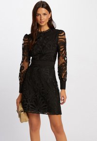 Robe en dentelle noire avec des motifs floraux délicats, longues manches transparentes et taille cintrée. Présente un col rond à bords festonnés et un tissu texturé.