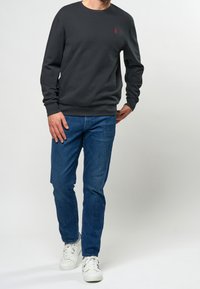 Mörkgrå sweatshirt med långa ärmar och rundad halsringning, prydd med en liten röd logotyp på bröstet. Bärs tillsammans med blå jeans och vita sneakers.