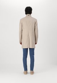 Abrigo de lana beige con cuello alto, corte recto y abertura en la parte trasera, combinado con vaqueros de denim azul y zapatos de color beige claro.