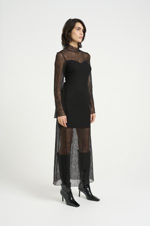 Vestido de encaje negro con mangas largas y superposición transparente; corsé ajustado, cuello alto y falda acampanada. Usado con botines brillantes negros.