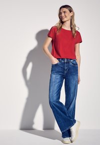 Maglietta rossa a maniche corte abbinata a jeans in denim blu a gamba larga. Sneakers bianche e un design semplice senza decorazioni o fantasie.