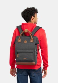 Zaino grigio con accentuazioni in pelle marrone, spallacci regolabili, manico superiore, tasca frontale con zip e tasche laterali in rete, indossato da una persona.