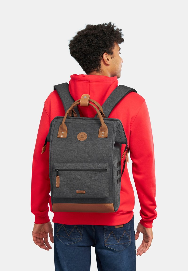 Zaino grigio con accentuazioni in pelle marrone, spallacci regolabili, manico superiore, tasca frontale con zip e tasche laterali in rete, indossato da una persona.