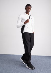 Balta garā piedurkņu polo krekls ar mazu logotipu, savienots ar melniem biksēm un melnbaltiem sporta apaviem. Modelis tur mazu melnu plecu somu.