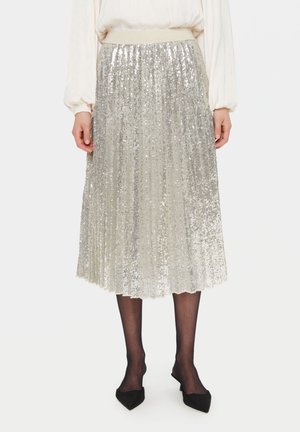 Srebrna plisowana spódnica midi z cekinami, noszona z przezroczystymi czarnymi rajstopami, czarnymi szpiczastymi balerinami i kremową bluzką z długim rękawem.