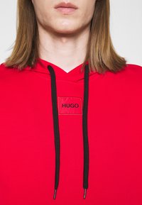 Czerwony hoodie z czarnym sznurkiem, z prostokątną naszywką z napisem "HUGO." Gładka struktura materiału, prosty design bez wzorów.