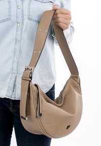 Isik hele teksariidest särgis, kes hoiab beežikat nahast poolkuukujulist käekotti laiendatava rihma ja lukuga.