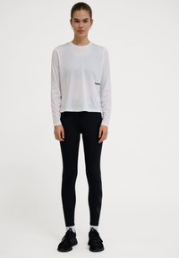 Weißes, langärmliges Sportoberteil mit bequemer Passform, kombiniert mit hochgeschnittenen schwarzen Leggings mit strukturierten Akzenten und Sportschuhen.