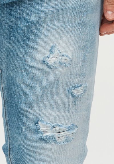 Ανοιχτό μπλε τζιν παντελόνι με πολλές φθαρμένες σκισίματα στην περιοχή του μηρού, που δείχνουν ξεφτισμένες λευκές κλωστές και ένα χέρι που ακουμπά κοντά.