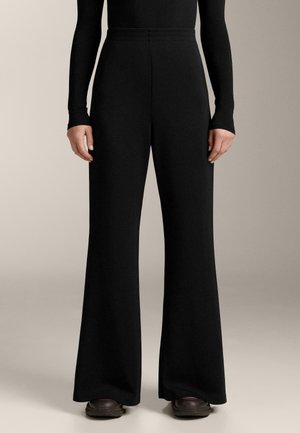 Person wearing high-waisted black wide-leg pants, black long-sleeve top, and dark sneakers, standing against a plain beige background.