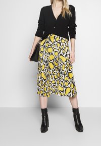 Czarny kardigan i żółta midi spódnica w panterkę. Spódnica ma czarne akcenty, zwiewny materiał oraz plisowany fason. Całość uzupełniają czarne botki za kostkę.