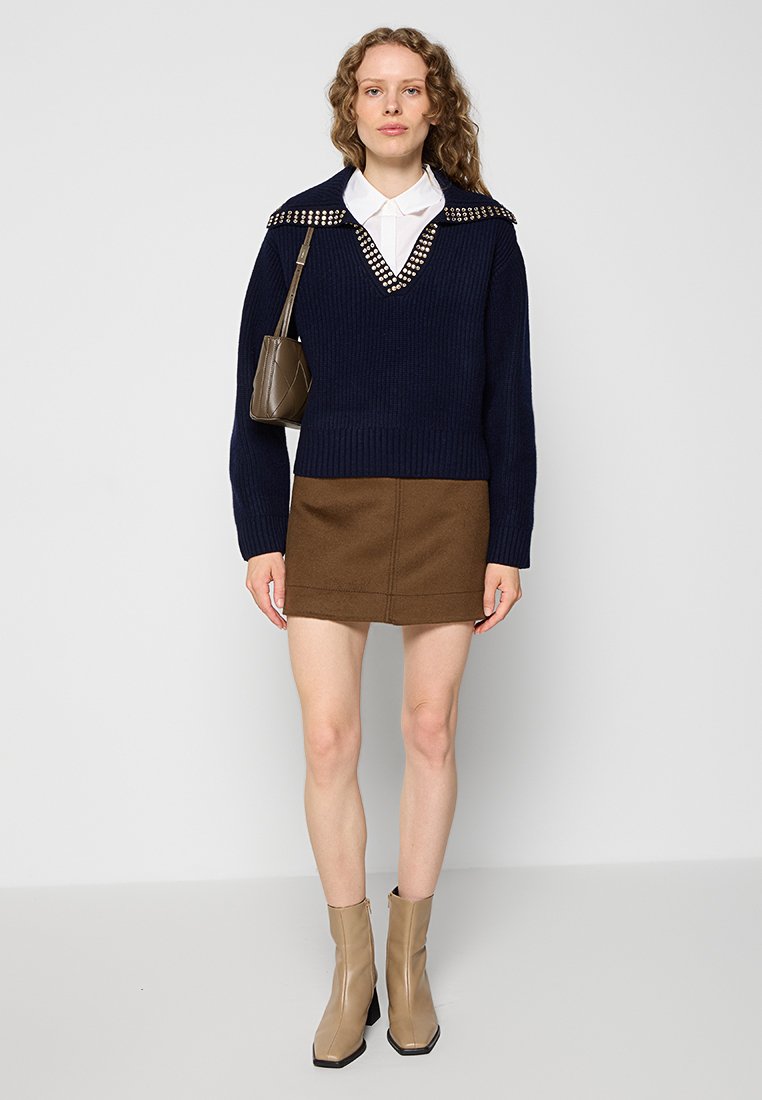 Pull en maille côtelée navy avec col clouté, mini jupe marron et bottines beige. Le col blanc contrastant avec le pull.