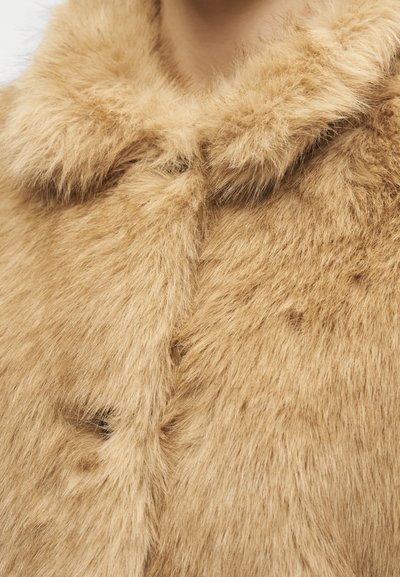 Gros plan sur le col d'un manteau en fourrure floue marron clair et la partie supérieure de la poitrine, montrant une texture douce et des fibres de fourrure denses.