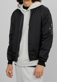 Svart bomberjacka i polyester, med dragkedja fram, ribbade manschetter och nederkant, har en sidficka med dragkedja, lagd över en grå hoodie.