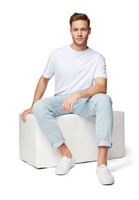 Jeune homme portant un t-shirt blanc, un jean bleu clair et des baskets blanches assis sur un bloc rectangulaire blanc sur fond blanc.