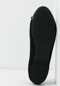 Chaussure noire à plat avec une tige en daim. La semelle en caoutchouc antidérapante présente une texture côtelée. Les détails sur le côté incluent un petit accent de bride.