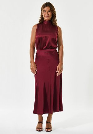Robe en satin bordeaux à col montant, jupe fluide et taille cintrée. Présente une fente pour faciliter les mouvements. Associée à des sandales à lanières.