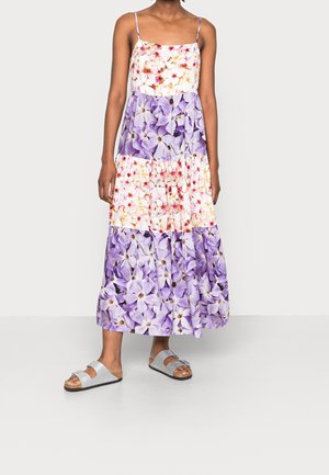 Robe maxi à bretelles spaghetti avec un design en étages, ornée de motifs blancs, roses et violets ; associée à des sandales grises.