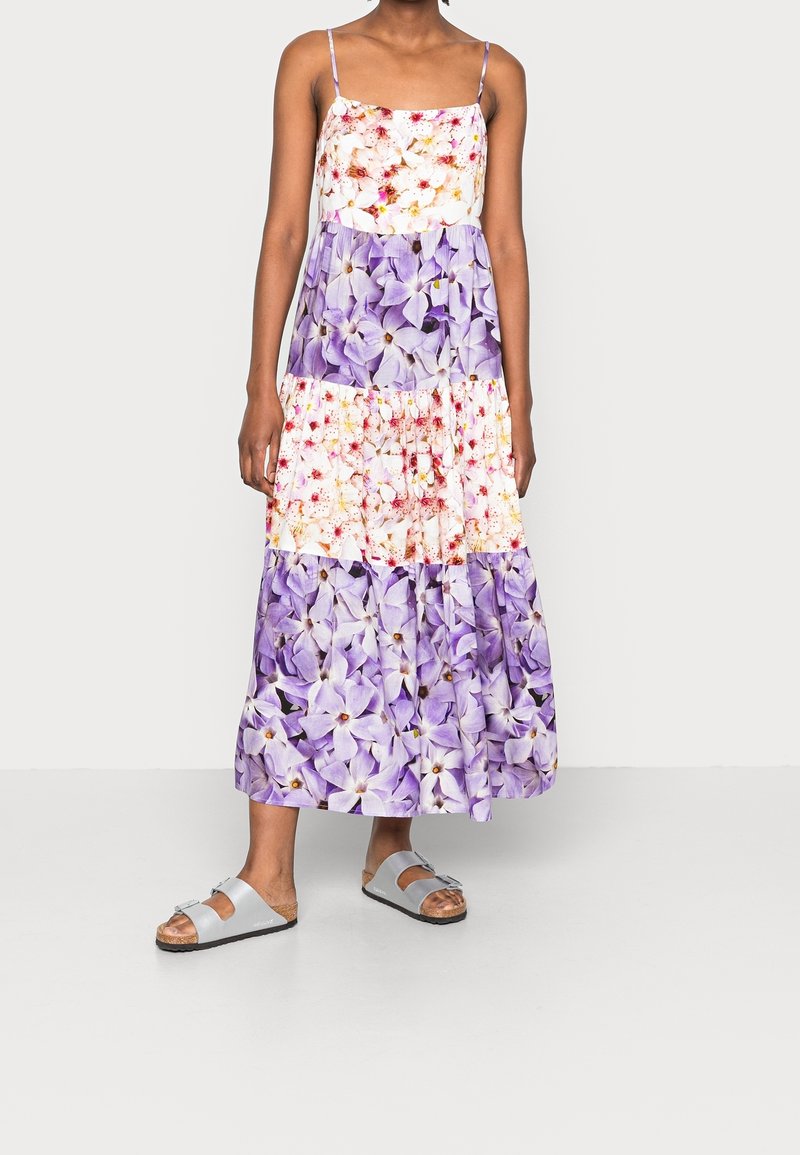Robe maxi à bretelles spaghetti avec un design en étages, ornée de motifs blancs, roses et violets ; associée à des sandales grises.